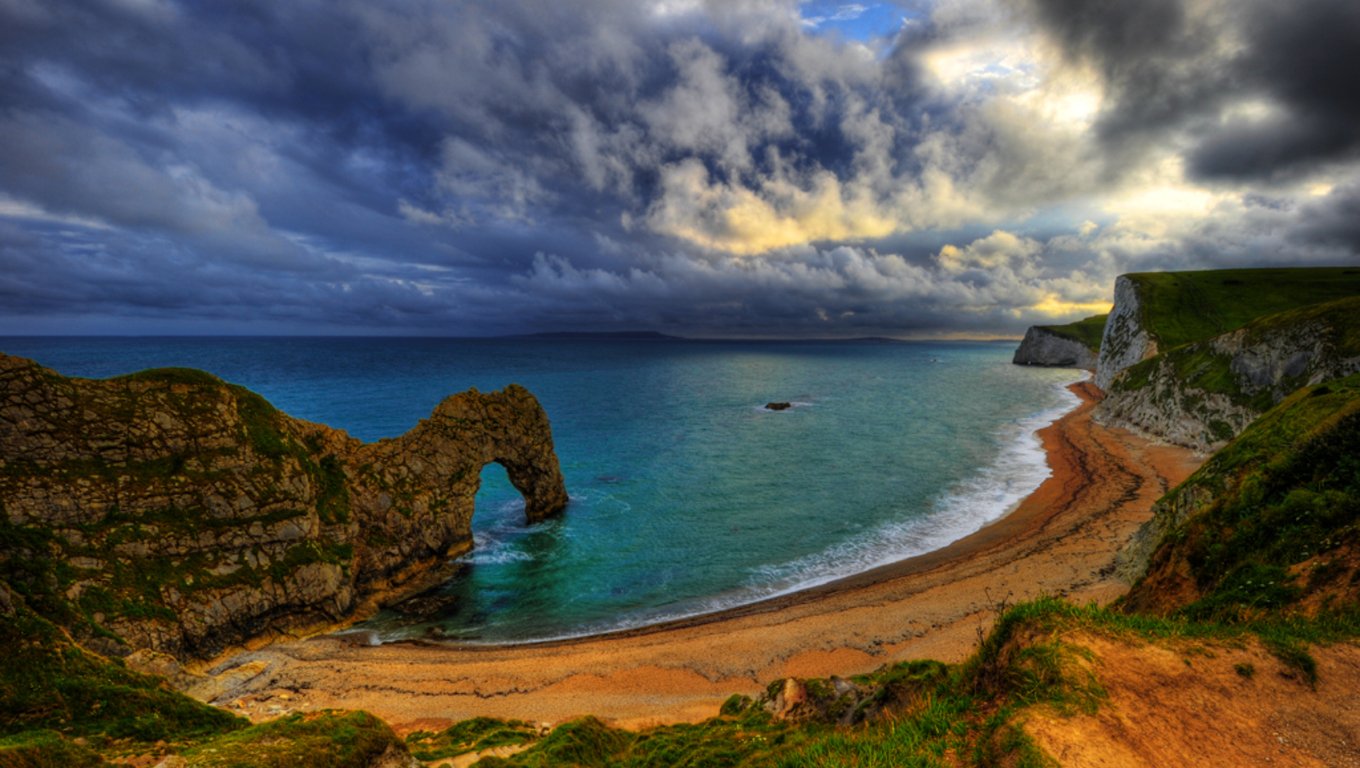 Durdle Door's natural limestone arch rises from the ocean beside a sandy beach under a dramatic, cloudy sky.