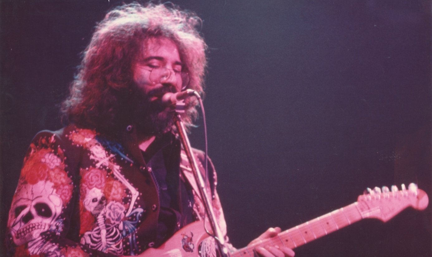 Grateful Dead guitarist passionately plays electric guitar on stage, wearing a vibrant jacket adorned with skull designs.