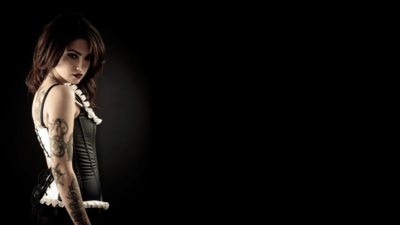 A woman with a striking tattoo poses confidently in a dark setting, wearing a stylish corset that highlights her unique character.