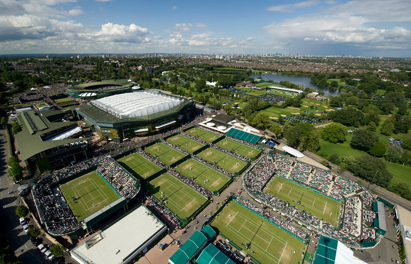 Aerial view of a large tennis complex with multiple grass courts, packed stands and surrounding greenery, capturing the energy of a sports tournament.