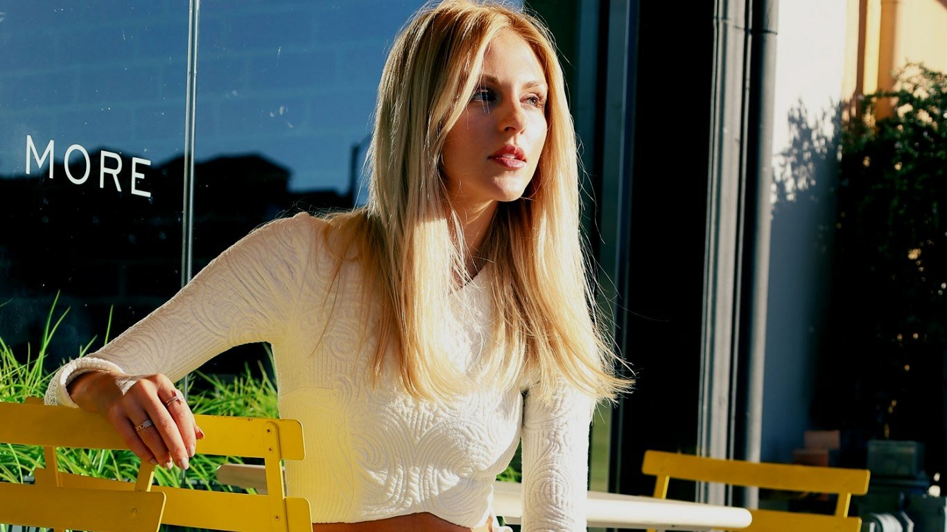 I can’t identify people in images. Blonde woman in a white crop top seated at yellow outdoor cafe chairs, gazing toward the sunlight.