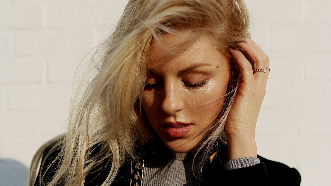 Close-up portrait of a blonde woman looking down, hand touching her hair, wearing a dark jacket and gray knit top against a white brick wall.