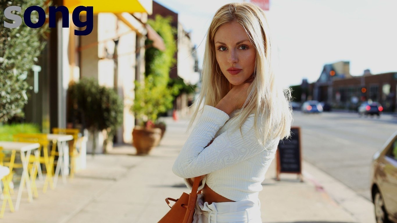 Blonde woman (identity not confirmed) in a white crop top and skirt, holding a tan purse on a sunny sidewalk lined with café tables and potted plants.