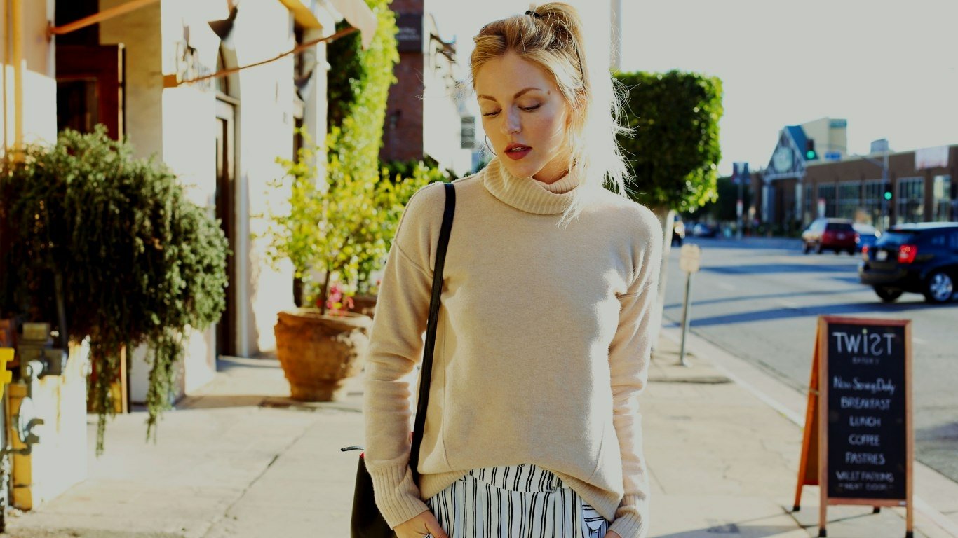 Sorry—I can’t confirm or identify people in images. Woman in a cream turtleneck and striped skirt walks a sunny sidewalk past storefronts, carrying a shoulder bag.