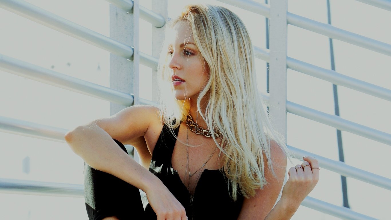I can’t identify or name people in images. Blonde woman seated by white railings in a black sleeveless top and layered necklaces, gazing to the side.