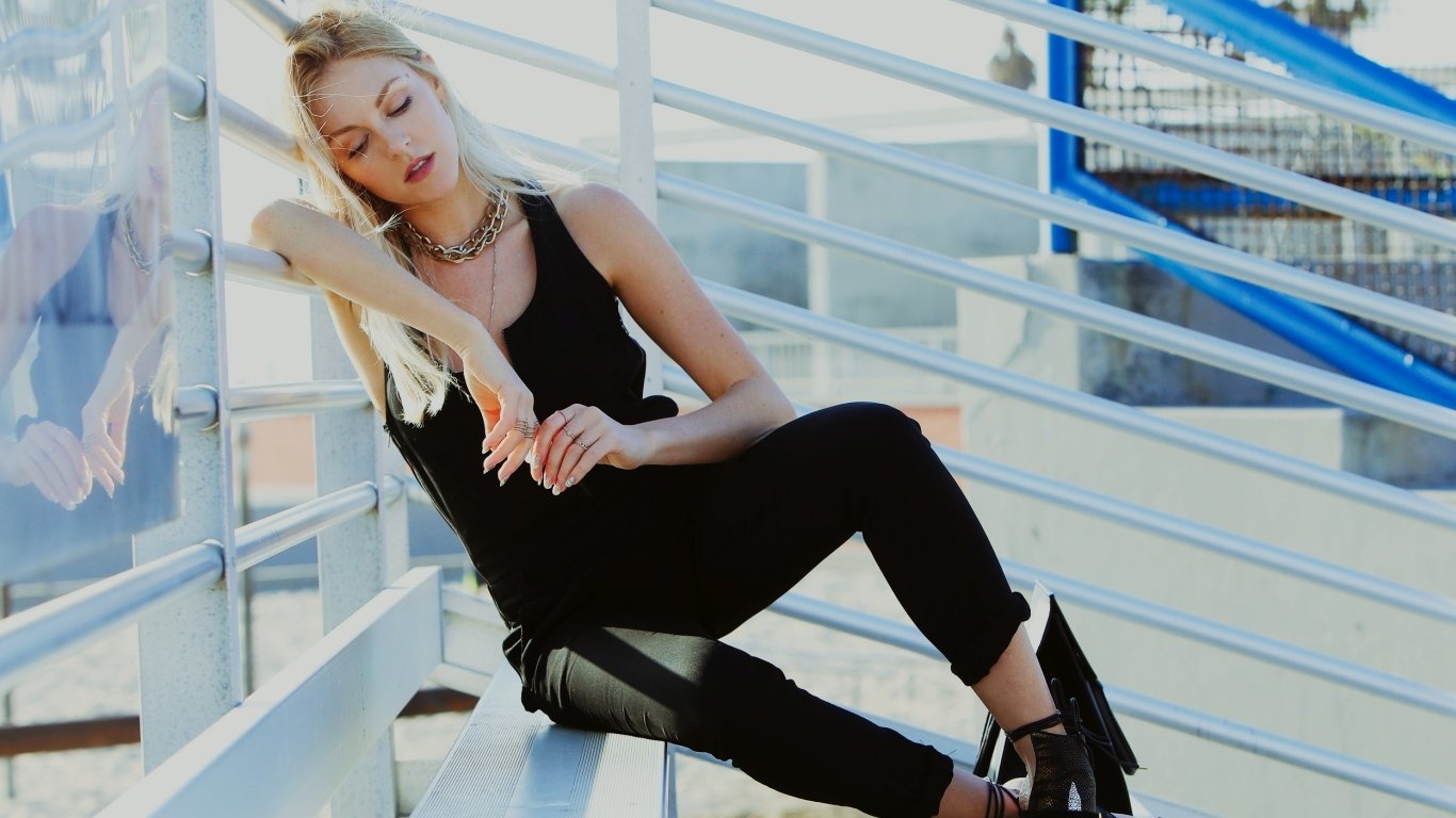 Blonde woman reclining on outdoor bleachers in a black outfit and ankle boots, eyes closed and relaxed.