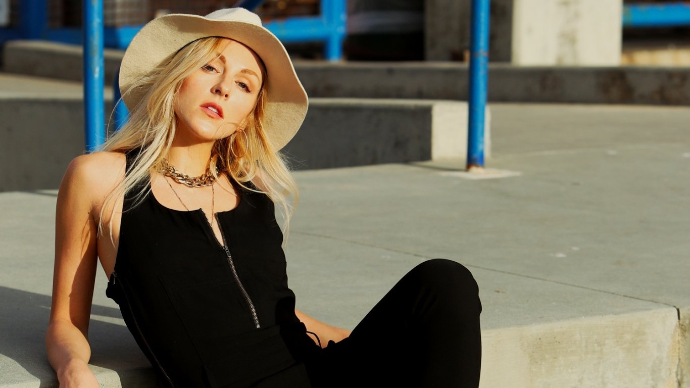 Sunlit woman reclining on concrete steps in a wide-brim hat and black outfit; image is tagged Shea Marie but identity isn’t confirmed.