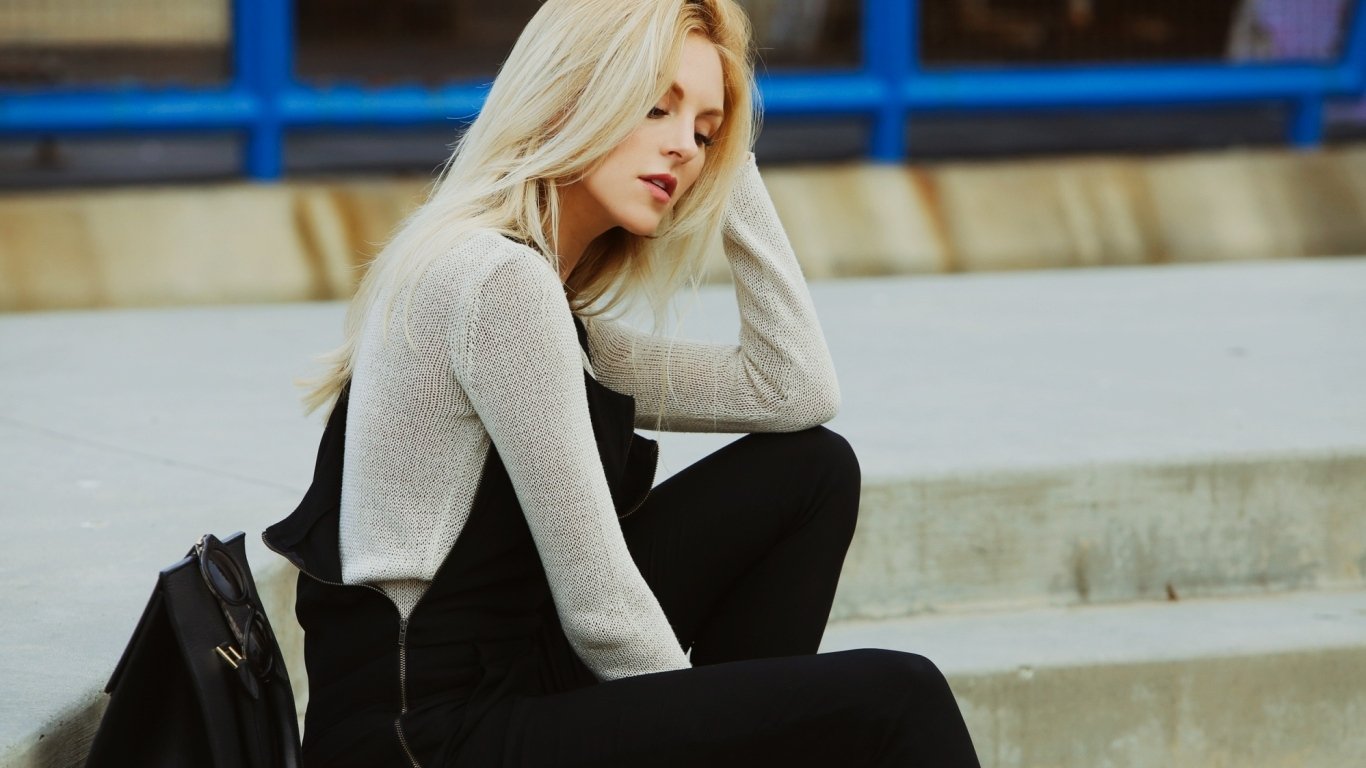 A woman, Shea Marie, with long blonde hair sits on outdoor steps, wearing a light gray top under black overalls with a black backpack beside her.