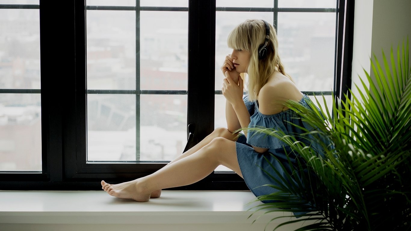 Woman named Lisa Dengler sits by a large window, wearing headphones and a blue off-shoulder dress, with a green plant nearby.