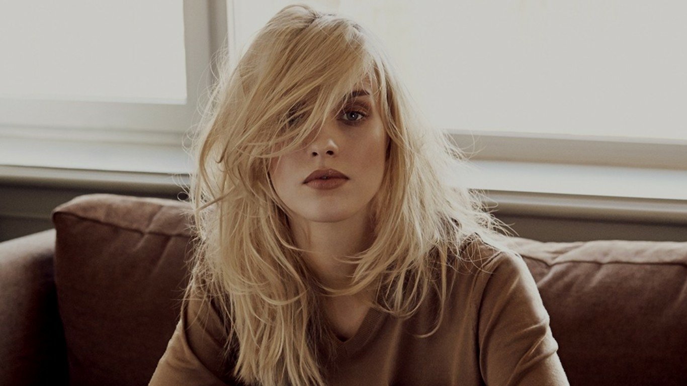 A woman with tousled blonde hair and a neutral expression sits on a brown couch near a window, embodying a relaxed and natural model look.
