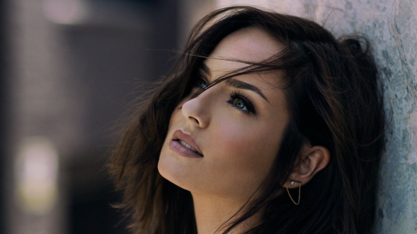 A woman model gazes upward, her hair softly tousled, showcasing a serene expression. The background features a blurred urban setting, adding depth to the portrait.