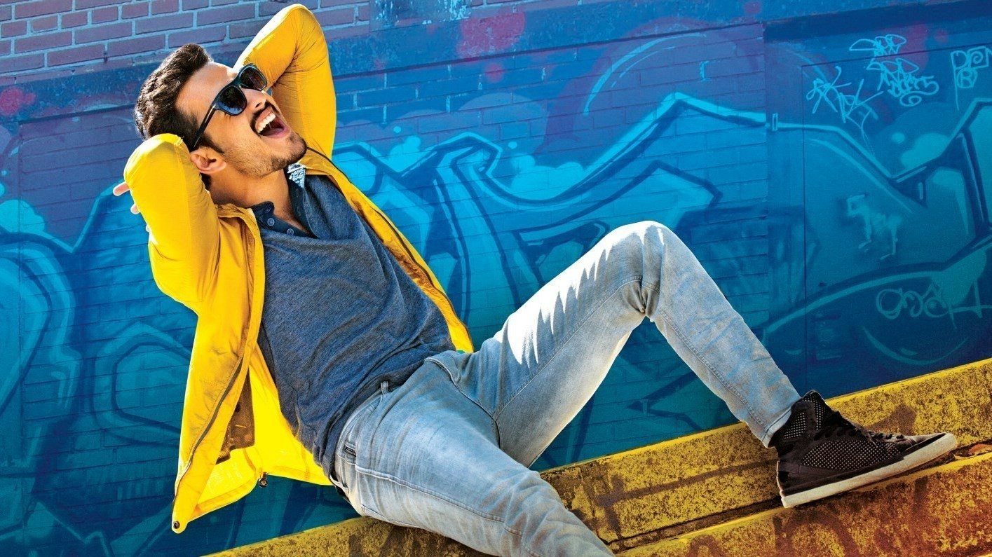 Akhil poses casually in a yellow jacket and sunglasses against a vibrant blue graffiti wall in a scene from the movie Akhil: The Power of Jua.