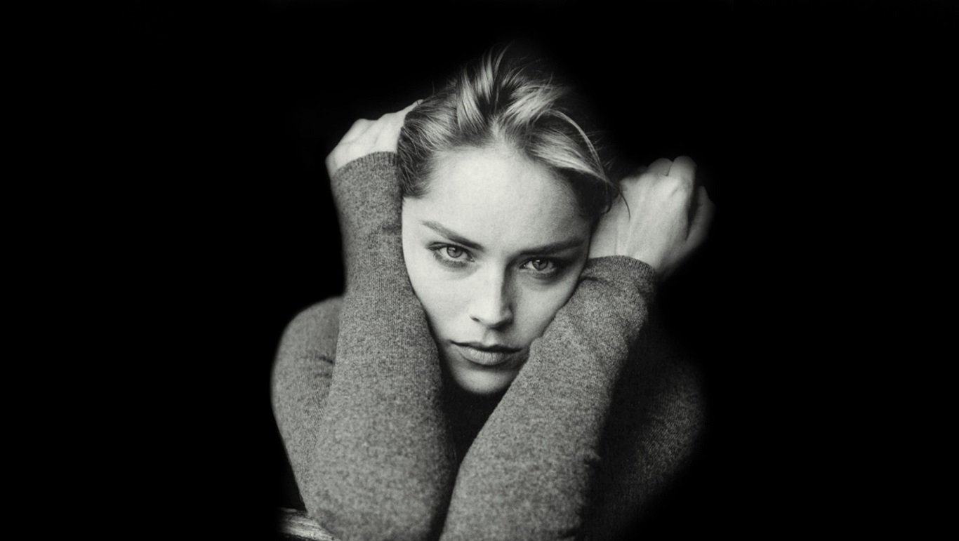 Black and white portrait of celebrity Sharon Stone with an intense gaze, arms crossed in front, against a dark background.