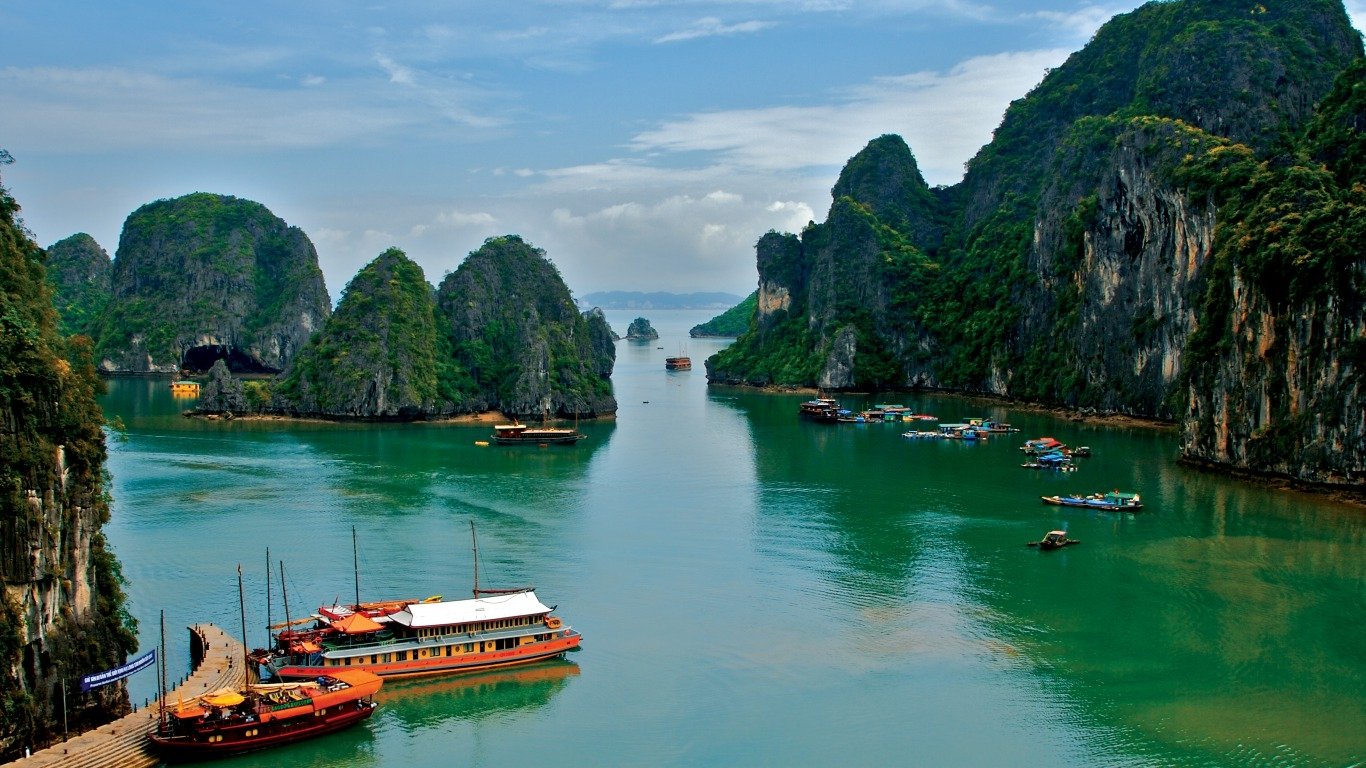 A serene seascape of Ha Long Bay, Vietnam, featuring traditional boats navigating between towering limestone islands under a partly cloudy sky.