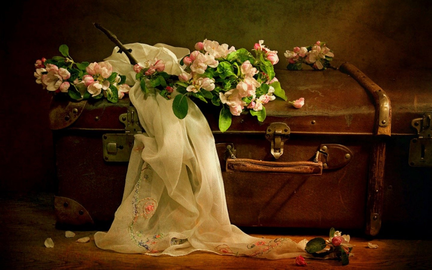 A vintage suitcase adorned with a delicate floral scarf and blooming pink flowers, captured in a warm, artistic still life photograph.