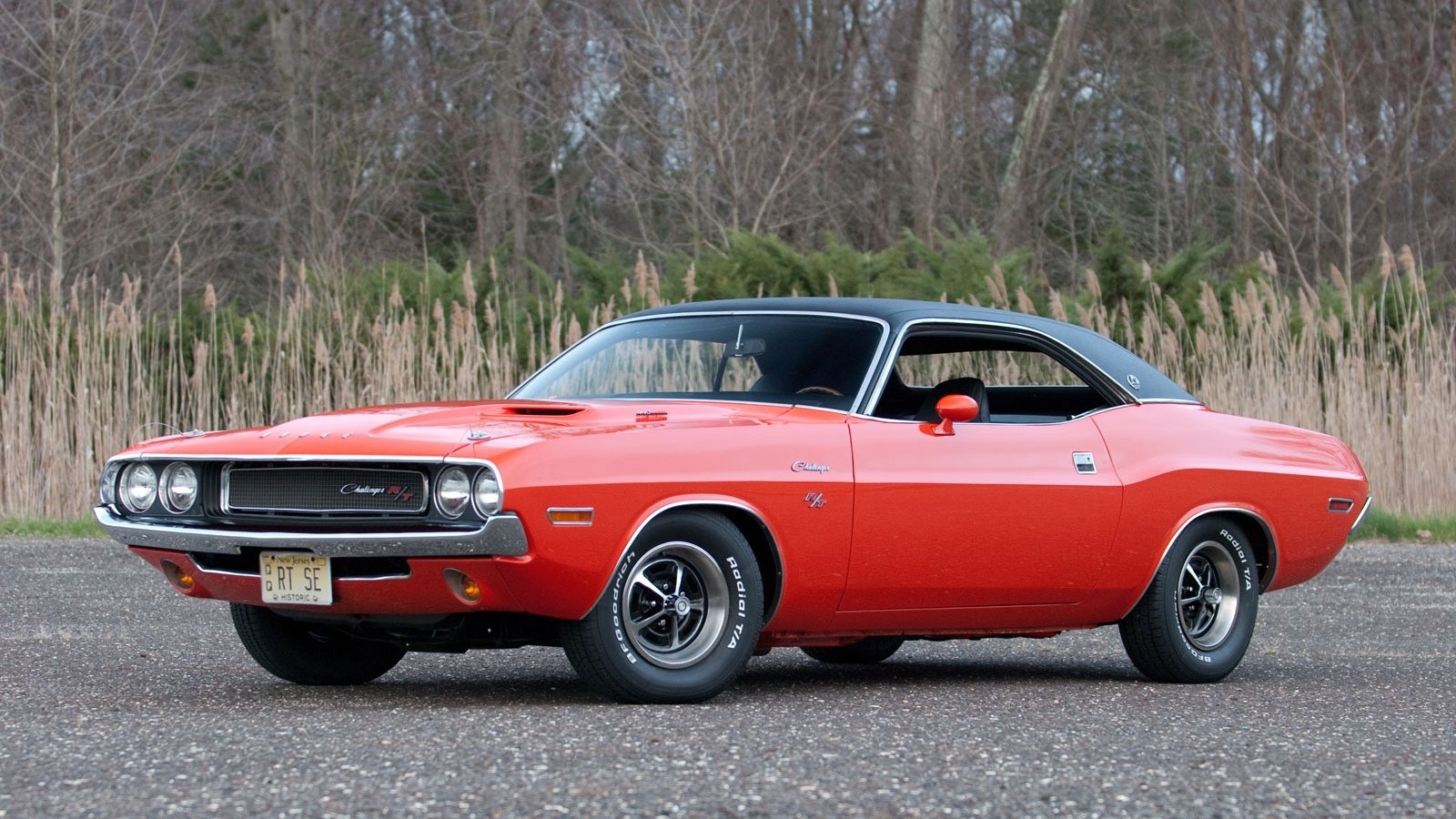 Bright orange Dodge vehicle — a classic muscle coupe with black vinyl roof and chrome rims — parked on pavement with trees and tall grass behind it.