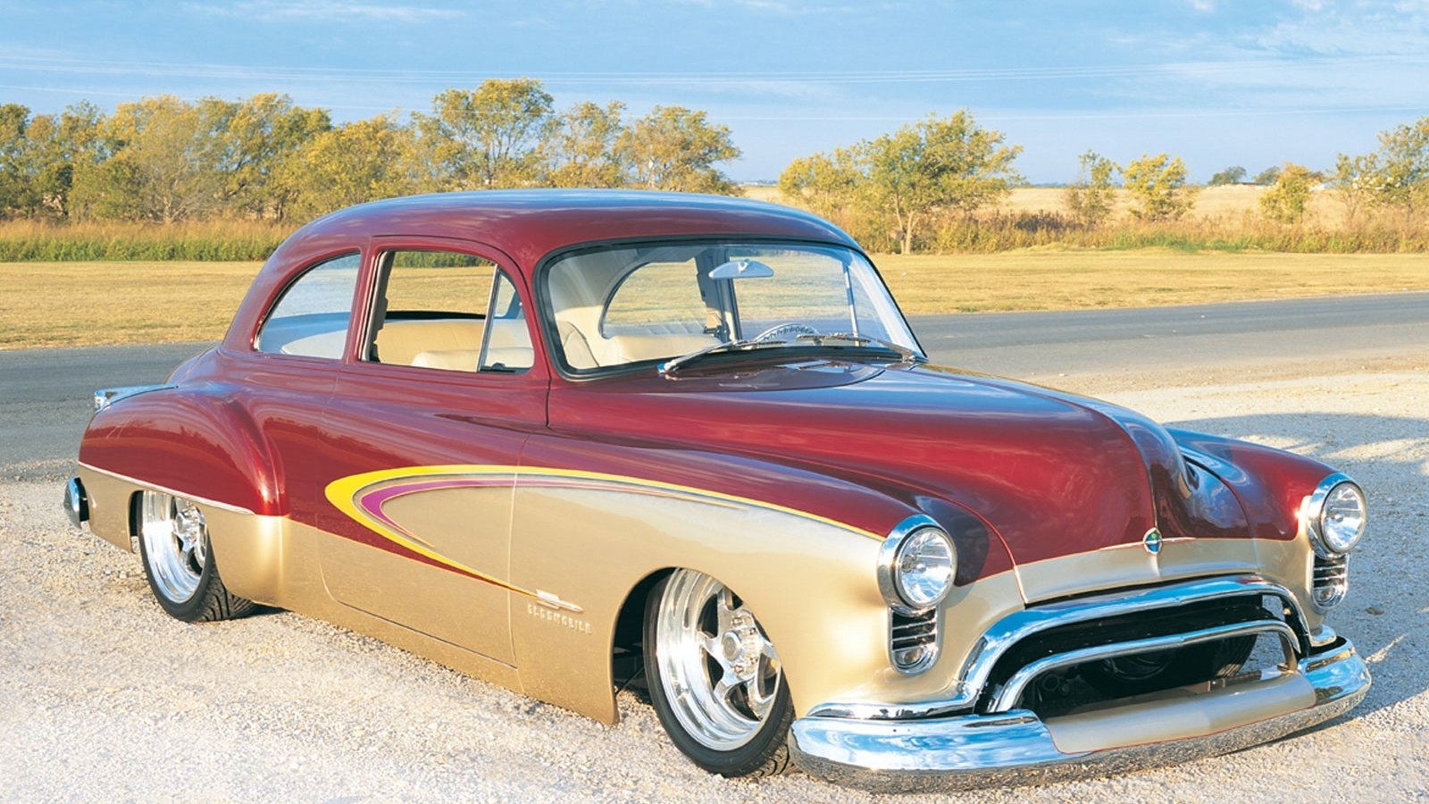 A vintage Oldsmobile vehicle with a two-tone red and gold paint job parked on a gravel road with a scenic backdrop of trees and open fields.