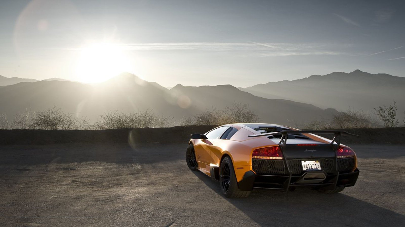 A Lamborghini Murcielago parked on a scenic overlook, facing the mountains, bathed in warm sunlight during sunset, showcasing its sleek design and vibrant orange color.
