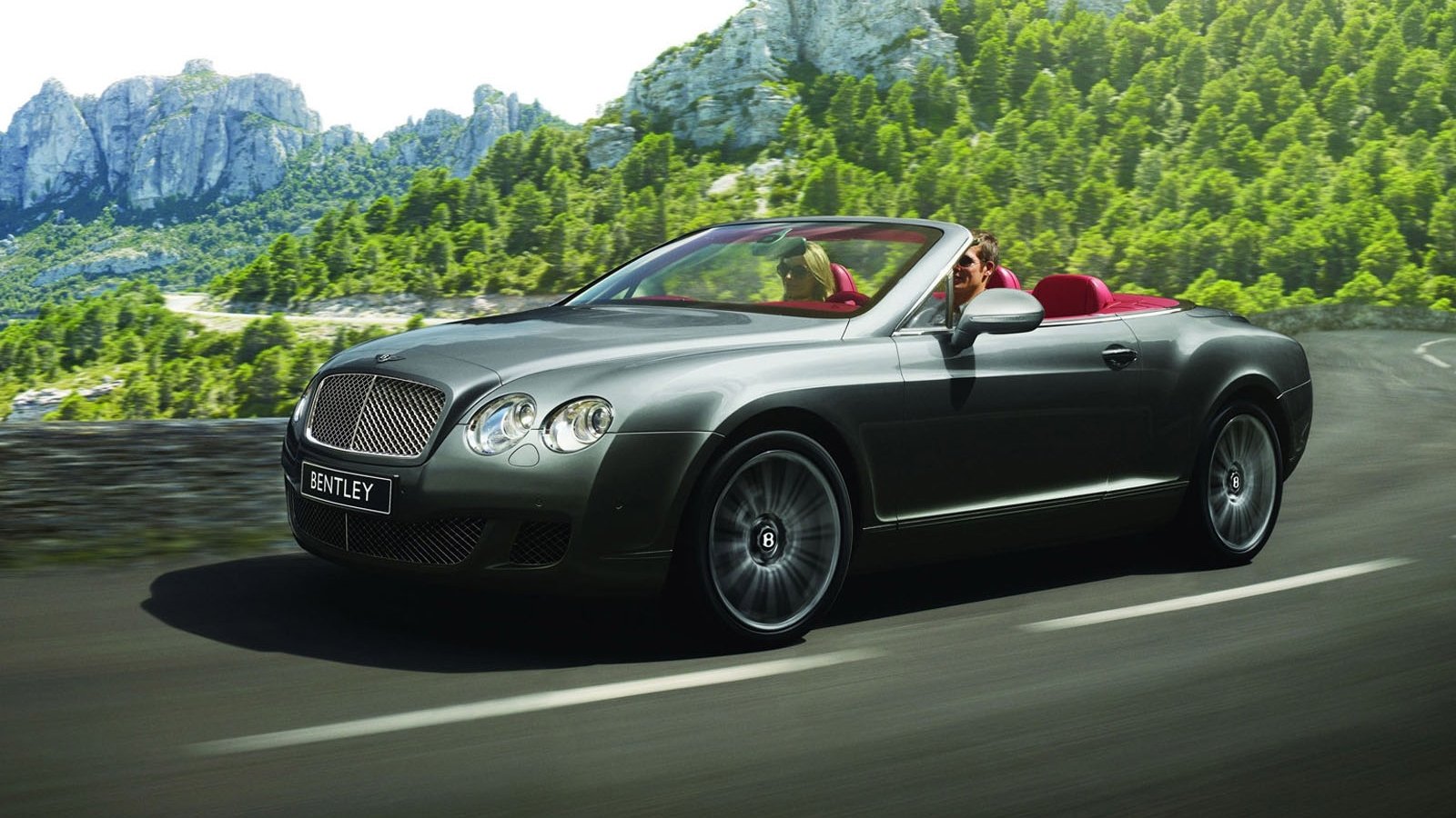 Silver Bentley convertible cruising on a winding mountain road, red leather interior visible against green hills and rocky cliffs.