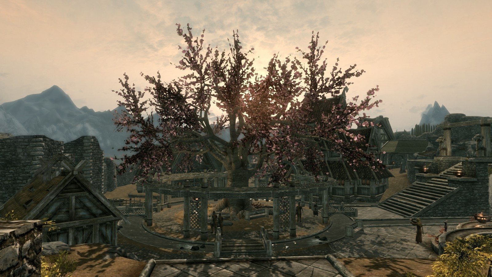 A scenic view of a village in The Elder Scrolls V: Skyrim at sunset, featuring medieval wooden buildings and a large tree in the center.