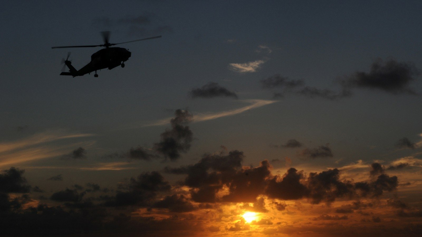 Silhouette of a military Sikorsky SH-60 Seahawk helicopter flying at sunset against a cloudy sky.