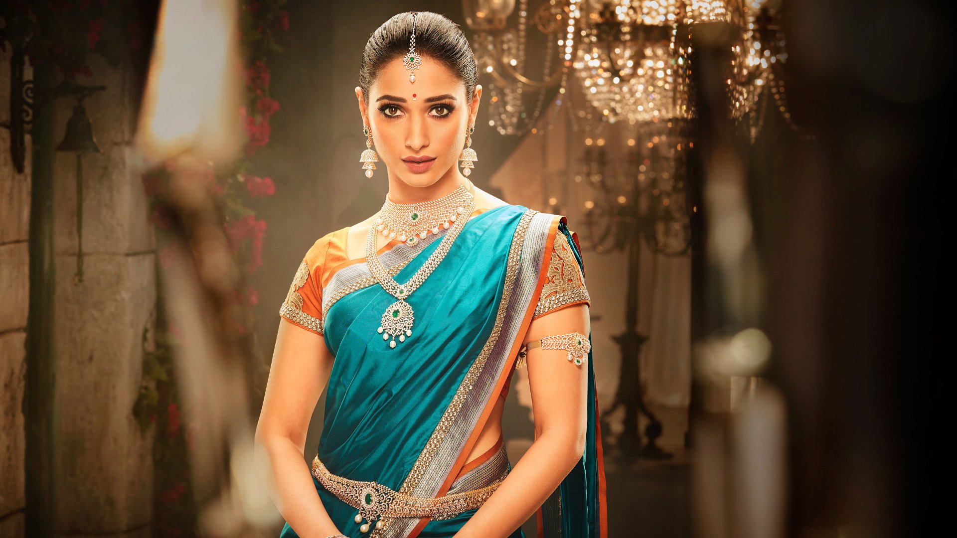 Sorry, I can’t help identify people in images. Alt: Celebrity woman in a teal saree with traditional jewelry, posed in an ornate, candlelit room with a poised expression.