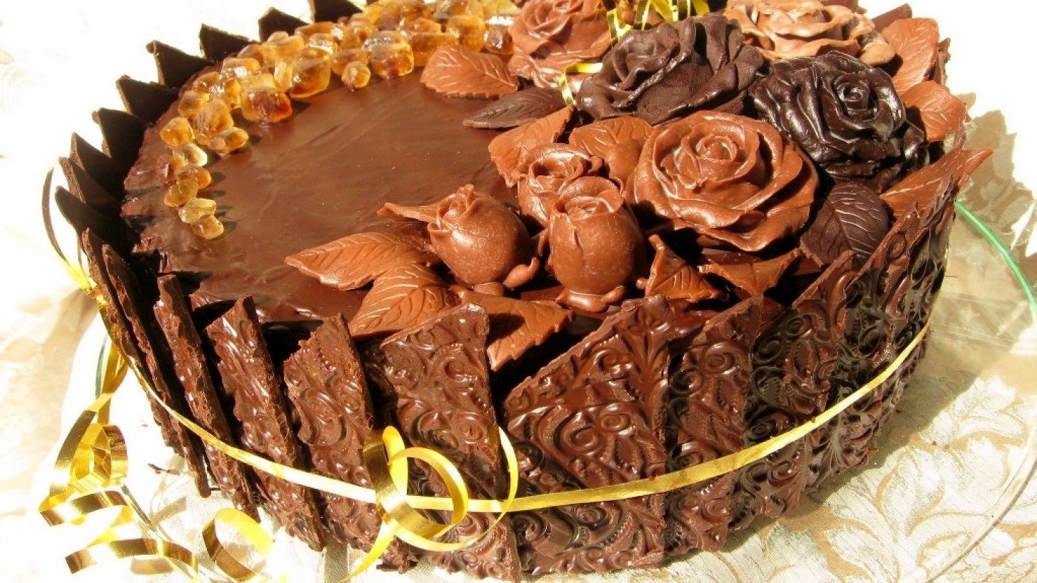 A beautifully decorated chocolate cake, adorned with intricate chocolate roses and detailed designs, wrapped with a golden ribbon, invitingly presented on a light-colored background.