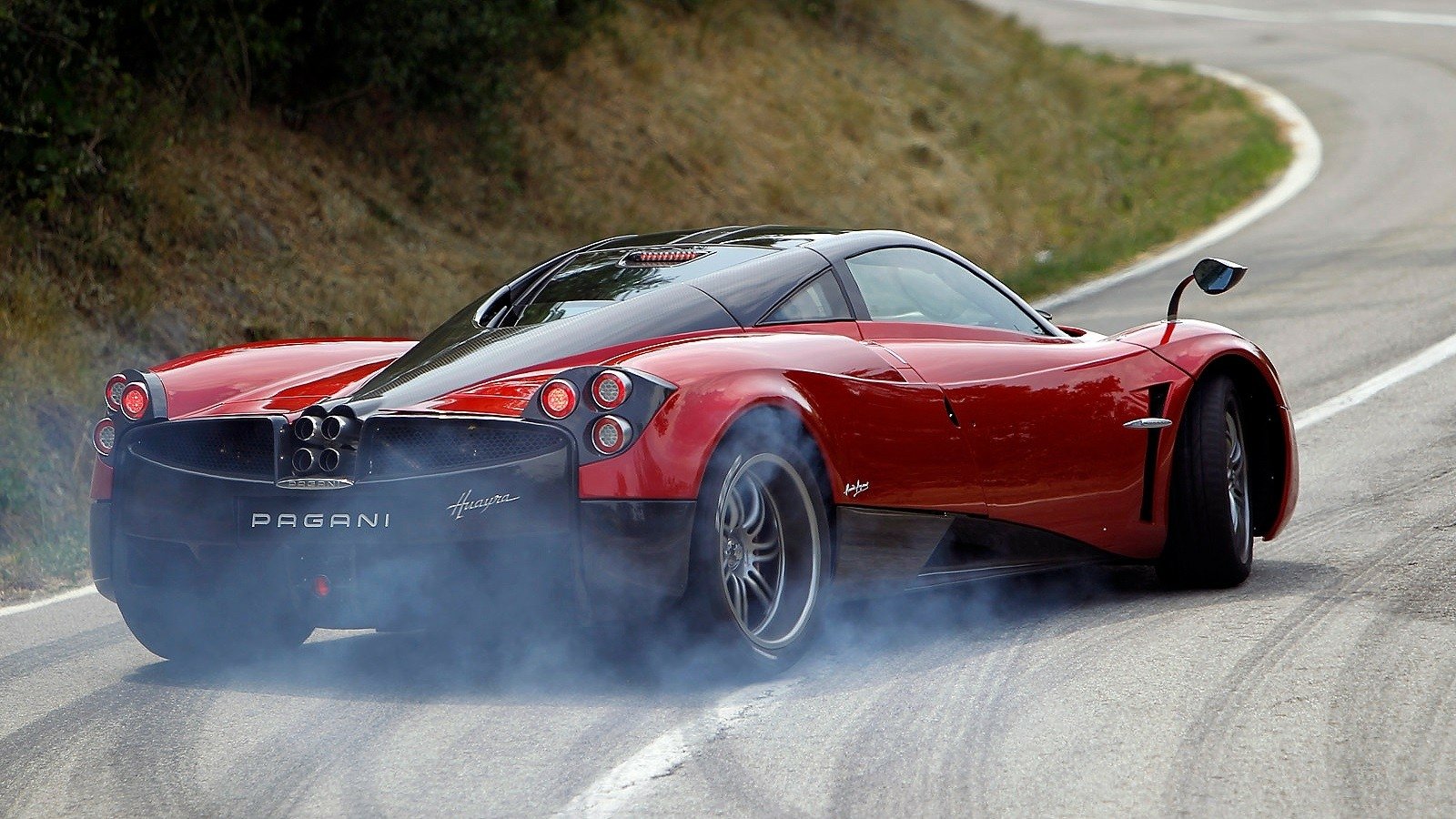 A striking red Pagani Huayra drifts around a curve, leaving a trail of smoke behind as it showcases its powerful performance on a winding road.