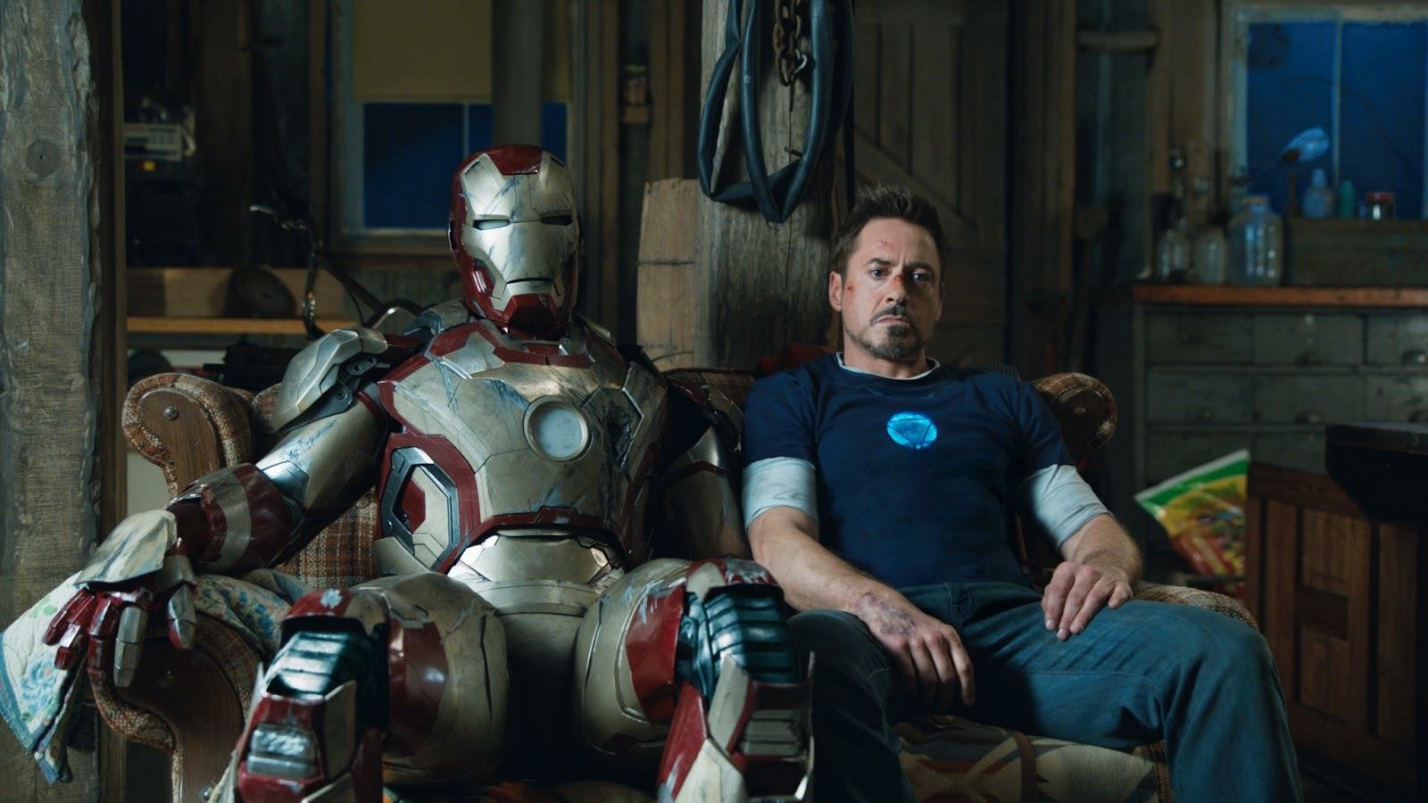 Tony Stark sits beside his Iron Man suit in a dimly lit workshop, a scene from the movie Iron Man 3.