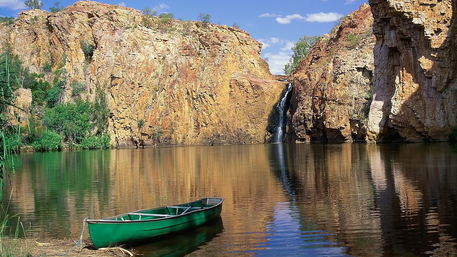 A green boat floats on calm water near rocky cliffs with a waterfall under a clear blue sky.