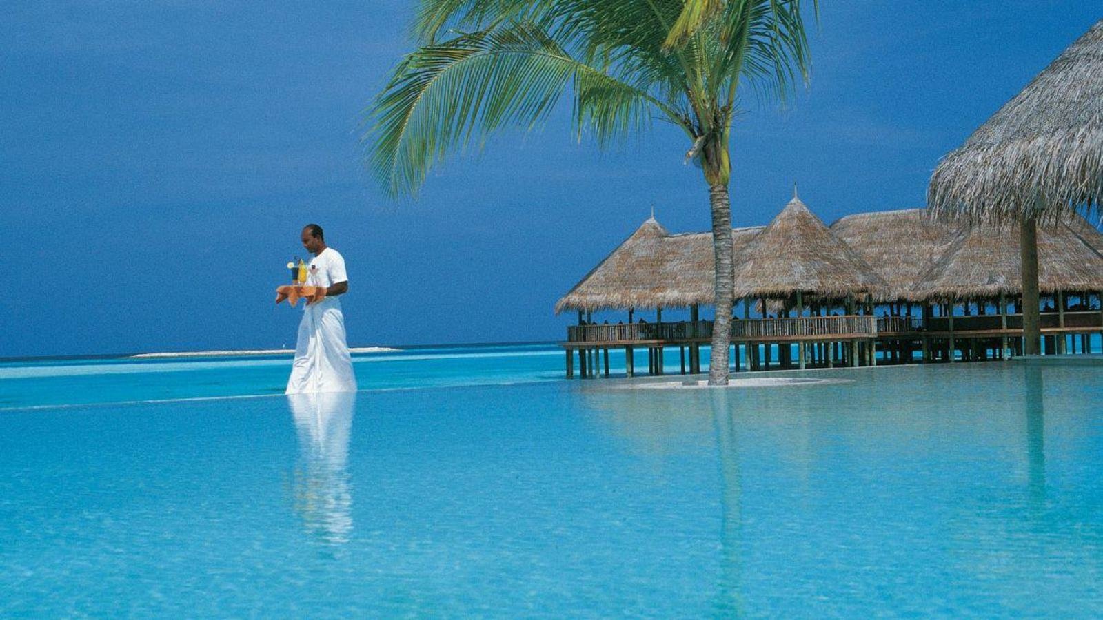 A person stands in clear, shallow ocean water near a palm tree and thatched-roof huts on a peaceful beach in the Maldives during a serene holiday.
