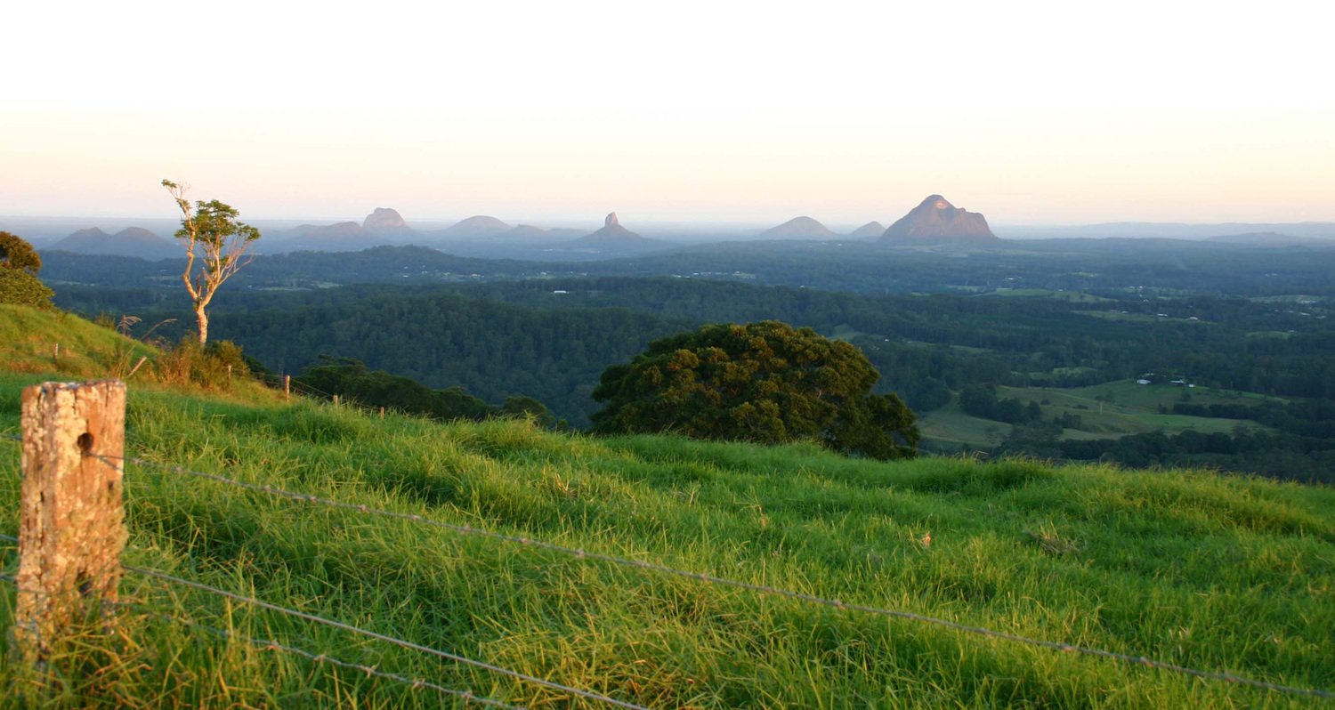 Lush green hillside with a fence post in the foreground, overlooking the distant Glass House Mountains and rolling valleys beneath a soft morning sky.