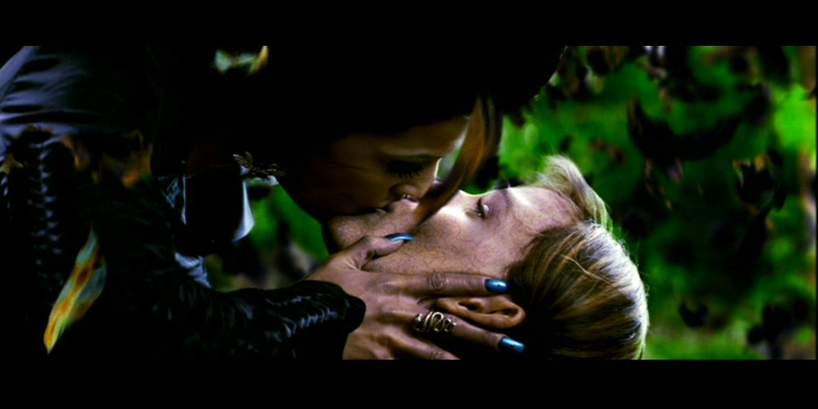 Close-up from the movie Elektra: a dark-clad woman leans over another, cupping her face in a tense, intimate moment amid a shadowy, leafy backdrop.