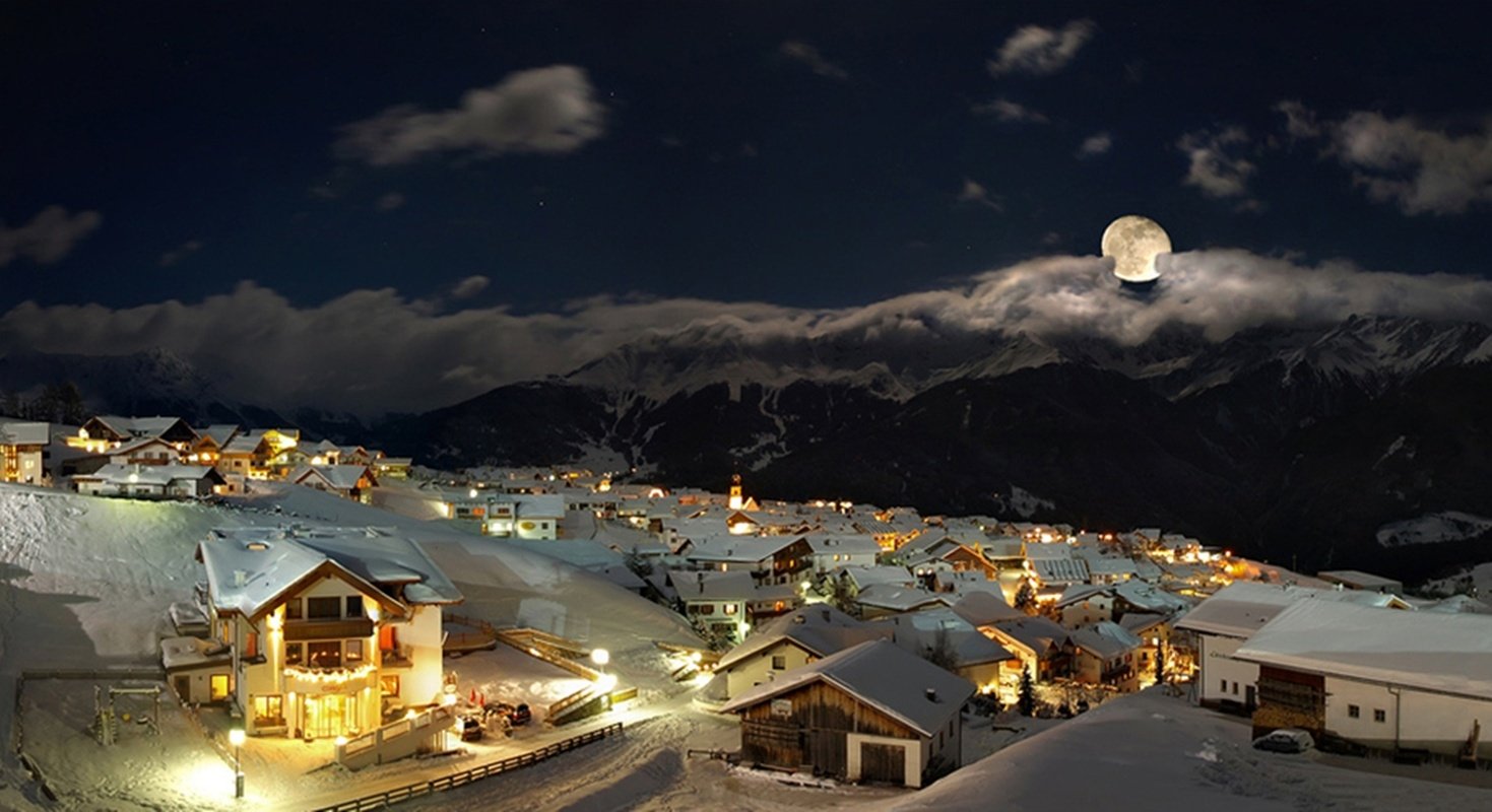 A man-made village covered in snow, illuminated by warm lights under a full moon and cloudy night sky with mountains in the background.