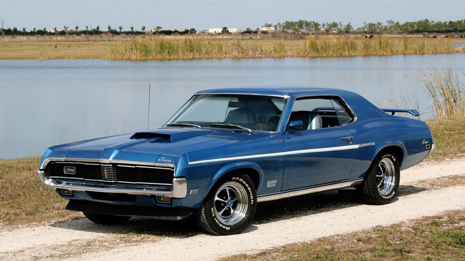 Blue Mercury Cougar Eliminator vehicle parked on a gravel road beside a lake, chrome wheels, hood scoops and vinyl roof visible.