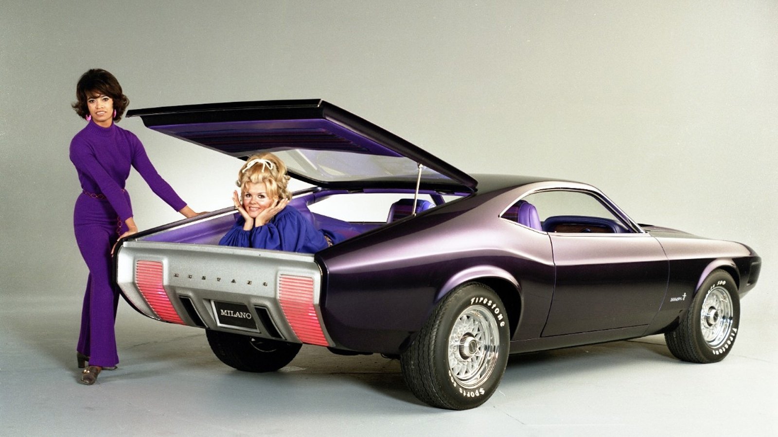 1970 Ford Mustang Milano concept vehicle — purple coupe with raised gullwing-like rear hatch, chrome wheels and dual tail lights, two people posing beside the open rear.