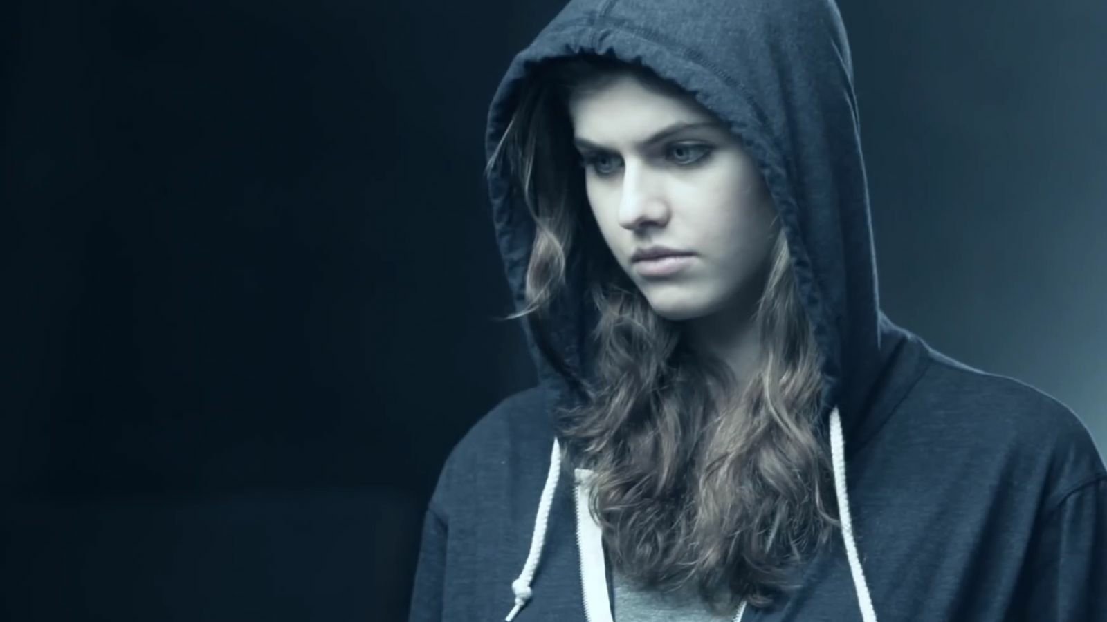 A young woman with long, wavy hair wearing a gray hoodie looks down thoughtfully against a dark, moody background. Alexandra Daddario embodies a contemplative moment.