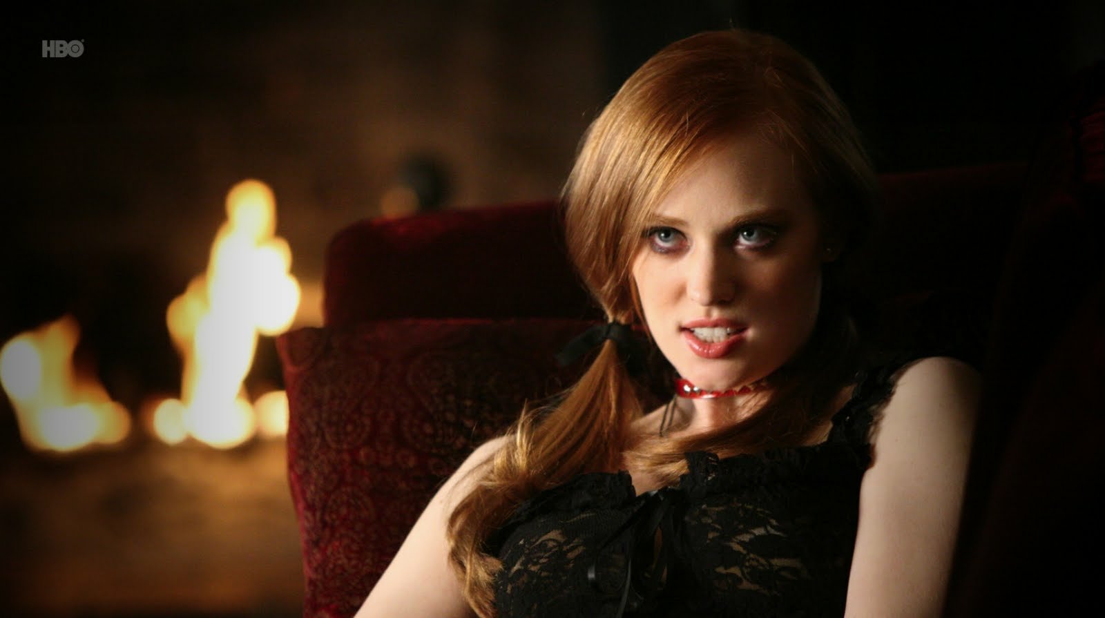 A scene from the TV show True Blood featuring a woman with red hair and intense eyes sitting near a glowing fireplace.