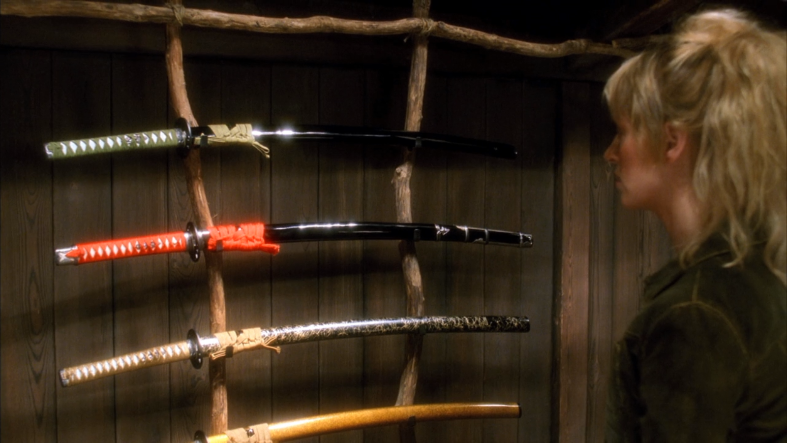 A scene from Kill Bill: Vol. 1 showing a woman looking at a rack of four samurai swords mounted on a wooden wall.