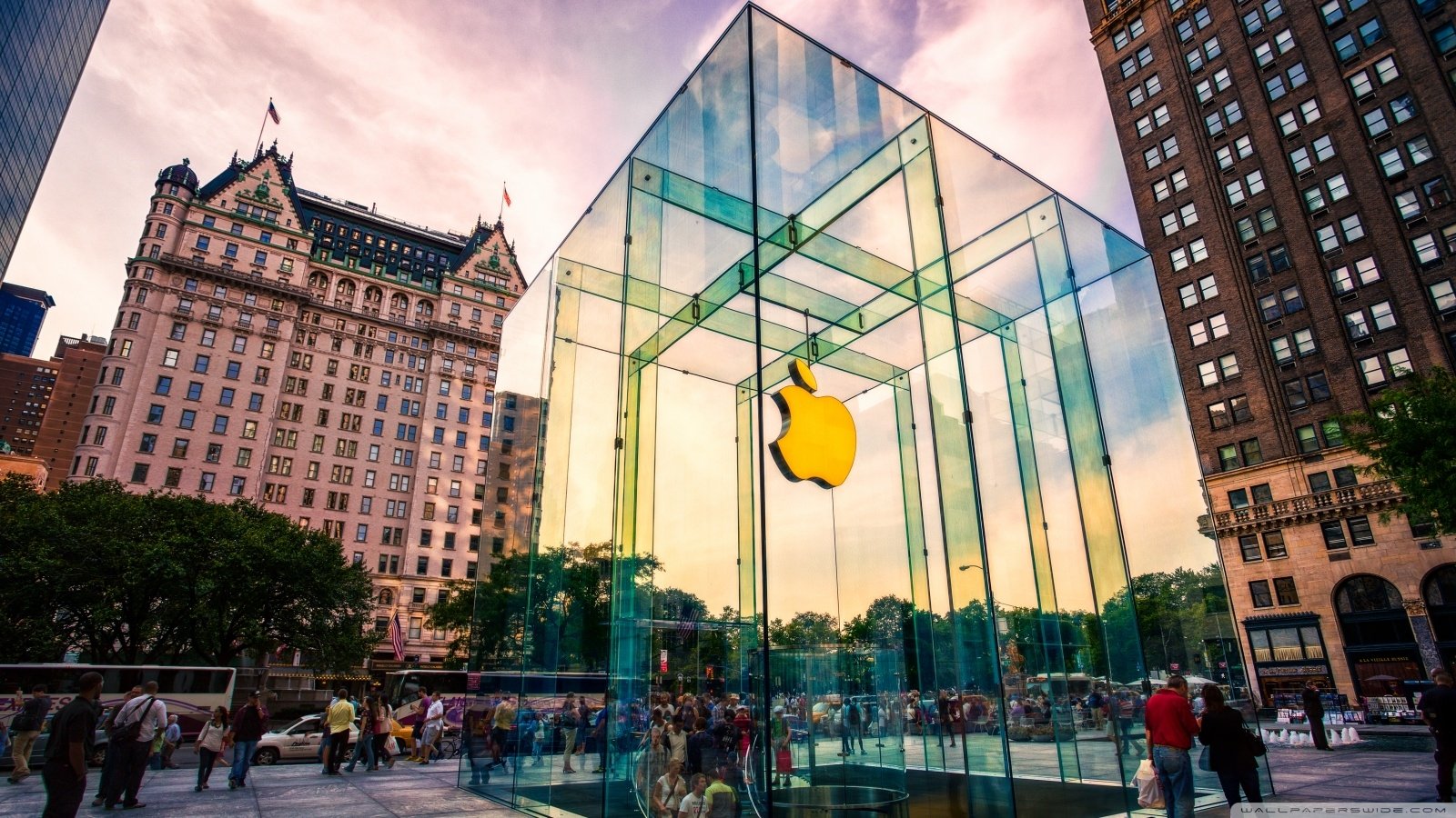 Glass-cube Apple Inc. store with glowing Apple logo set among man-made city buildings, pedestrians milling in the plaza.