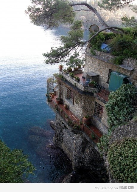Man-made stone building: a coastal house perched on a rocky waterfront cliff with terraced balconies, potted plants and trees overlooking the calm blue sea.