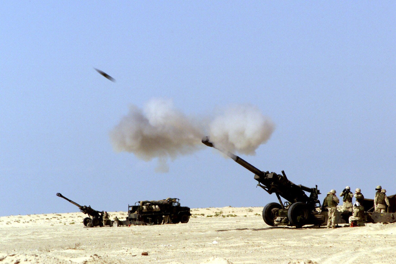 A soldier operates artillery in a desert landscape, as a shell is fired into the sky, surrounded by fellow military personnel and equipment. Smoke billows from the cannon's muzzle.