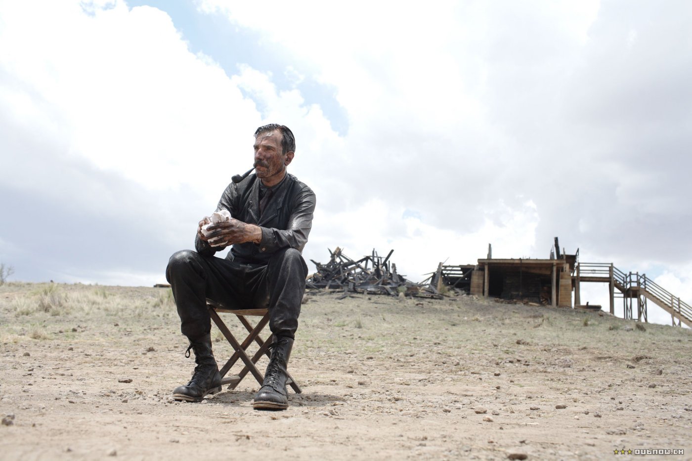 Man in period clothes sits on a folding chair in a dusty oilfield, bottle in hand, rig and cloudy sky behind — still from the movie There Will Be Blood.
