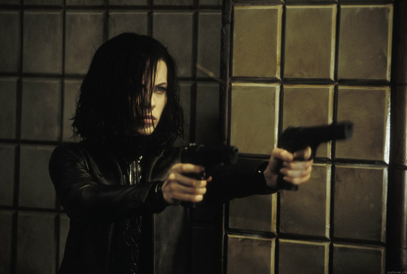 Kate Beckinsale as Selene in *Underworld: Evolution*, fiercely aiming dual pistols in a dimly lit setting with gritty, urban textures in the background.