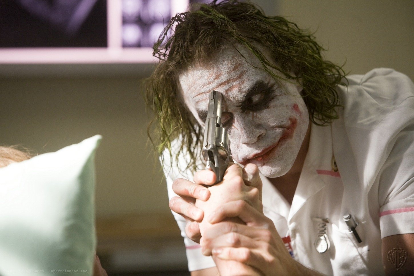 Heath Ledger as the Joker in *The Dark Knight*, featuring his iconic white makeup and manic expression, points a gun while interacting with another character in a tense scene.
