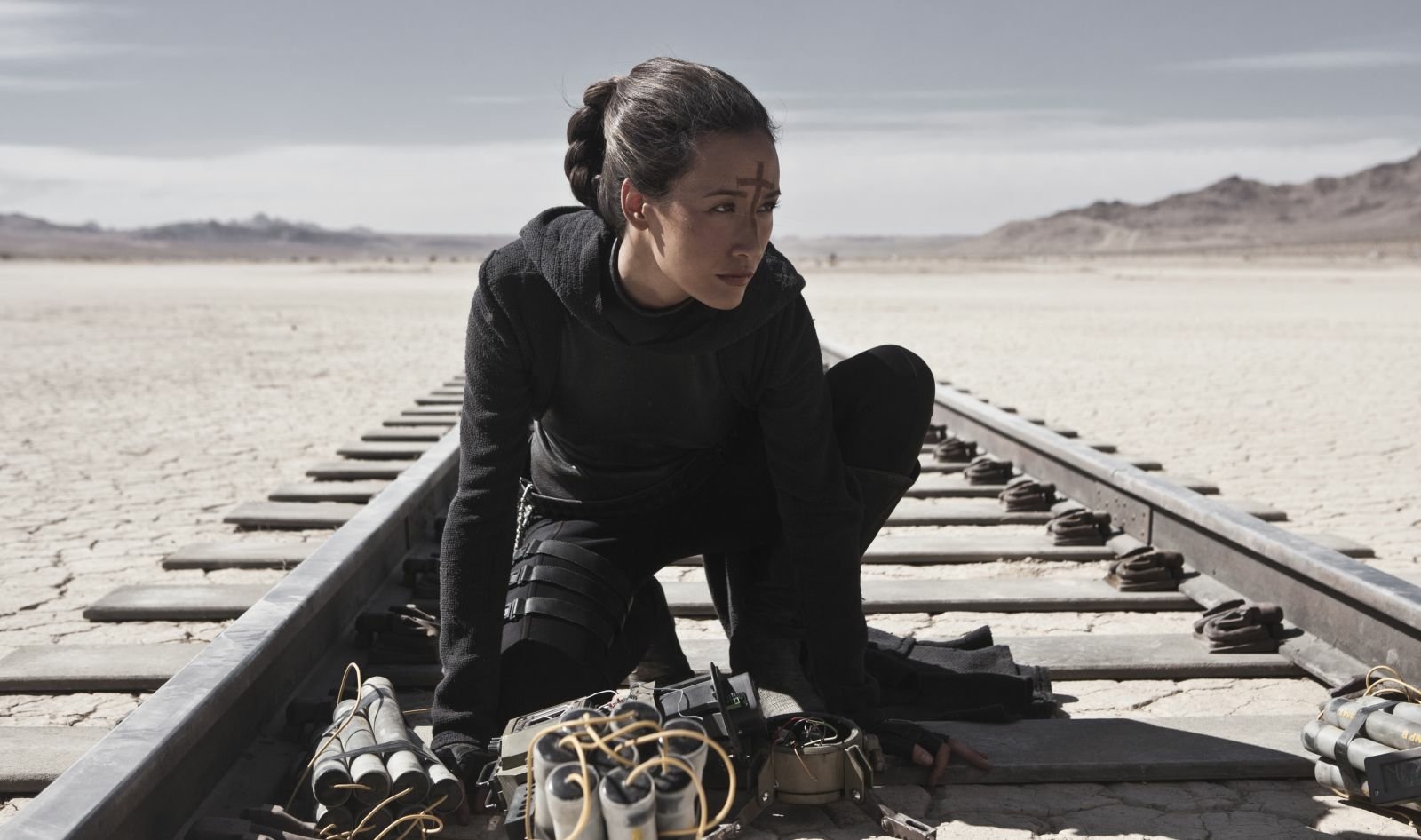 Maggie Q is depicted crouching on train tracks in a desolate landscape, surrounded by explosives, conveying a sense of tension and determination.
