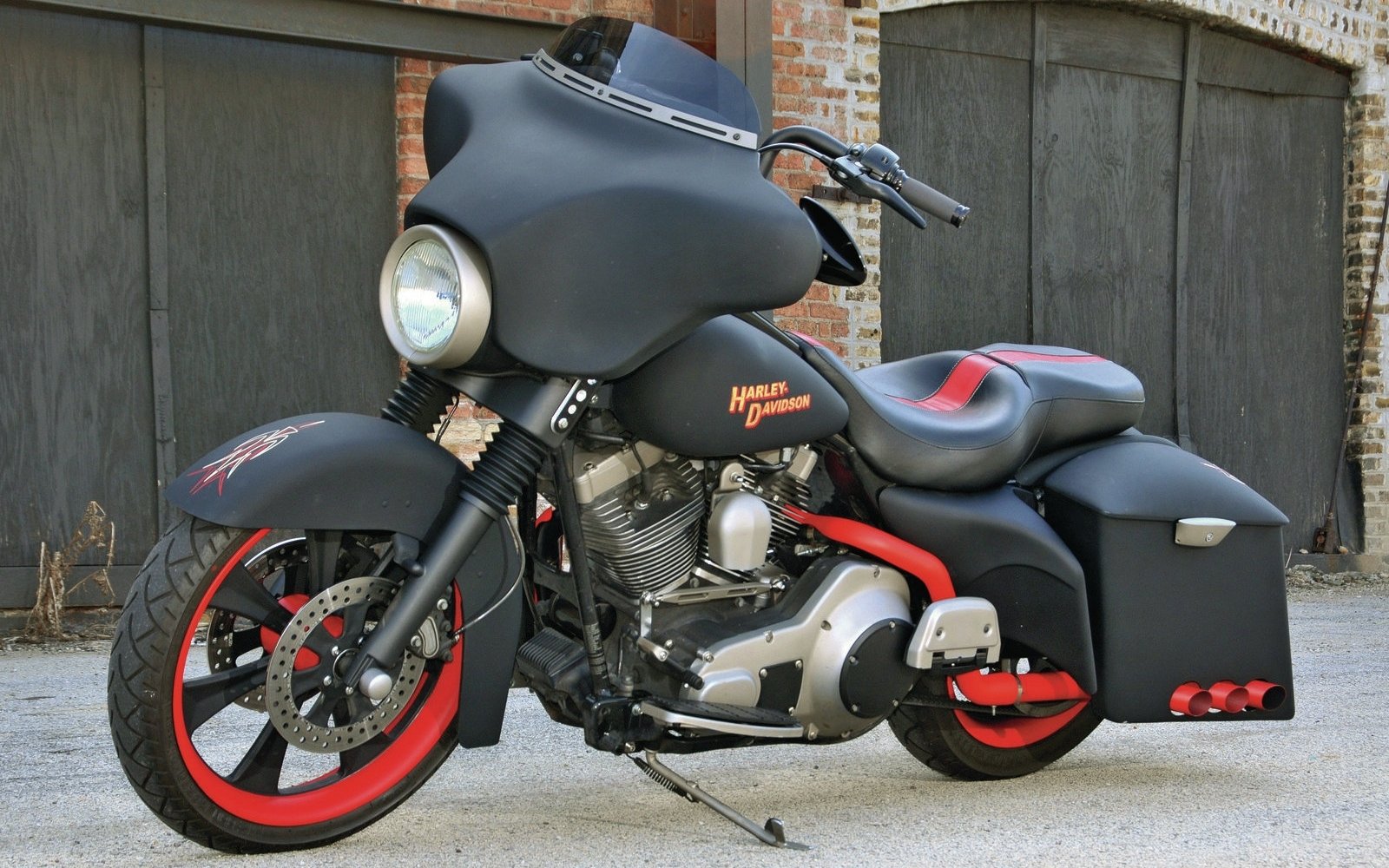 Custom Harley-Davidson motorcycle with matte black paint, red accents on wheels and engine, and hard saddlebags parked in front of brick garage doors.