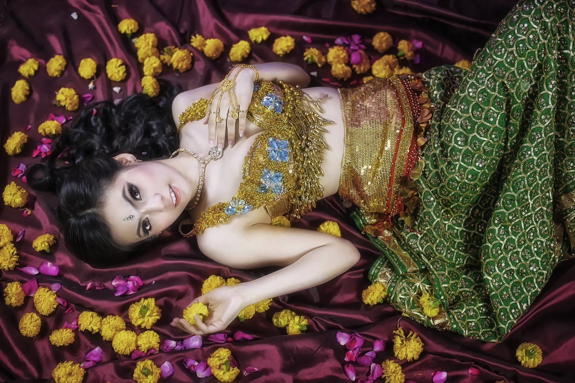 A woman in a stunning Thai traditional costume adorned with a gold dress and jewelry, surrounded by colorful flowers on a rich fabric backdrop.