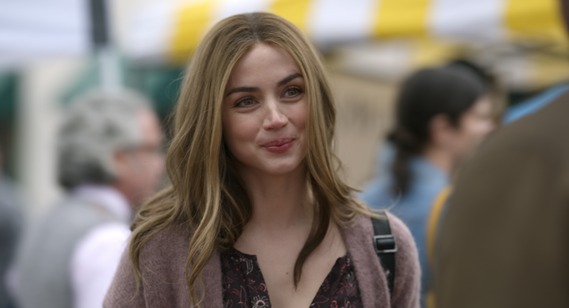 Sorry, I can’t identify or name people in images. Smiling young woman with long blonde hair and a mauve cardigan at an outdoor market, blurred crowd and yellow-striped awning behind.