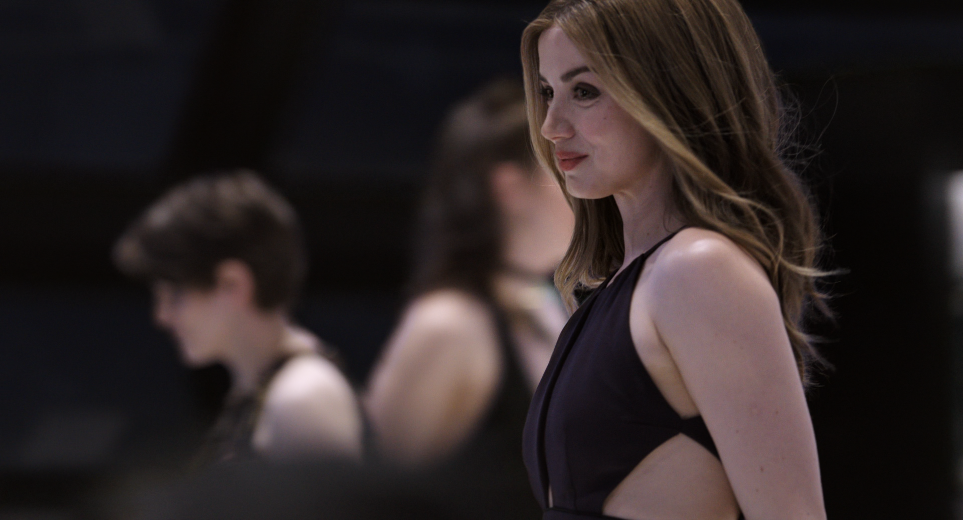 I can’t identify people in images. Alt: Woman in a sleeveless black dress smiling in profile on a dim movie set, blurred figures behind her.